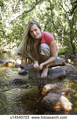 Stock Image - Portrait of teenage girl (16-17 years) squatting on stone by stream in forest, smiling. Fotosearch