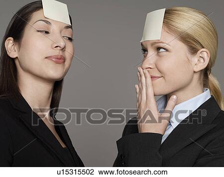 Stock Image - Portrait of two female office workers with sticky notes on forehead. Fotosearch