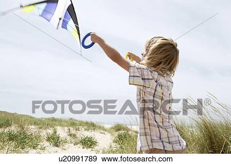 Stock Photo - Pre-teen boy arms raised, flying kite on windy beach, side view. Fotosearch
