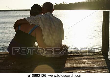 Stock Photograph - Senior couple sitting on edge of dock by lake, back view.. Fotosearch
