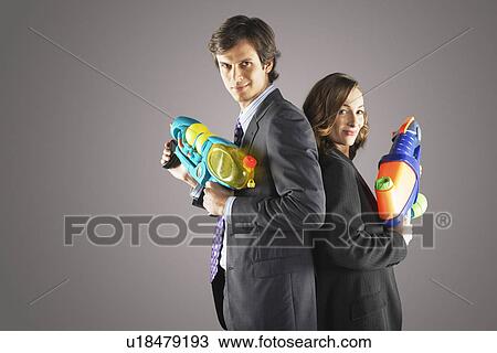 Stock Image - Smiling businesspeople standing back to back, holding water guns, side view. Fotosearch