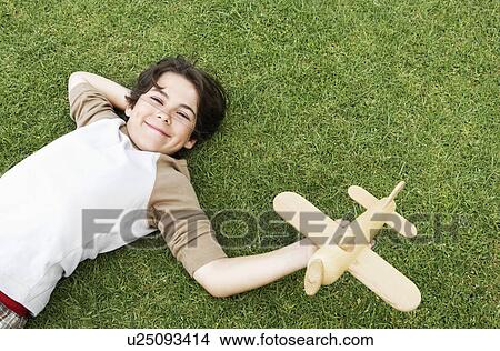 Picture - Smiling Pre-teen boy lying on back in grass, arm behind head, holding toy airplane, elevated view. Fotosearch