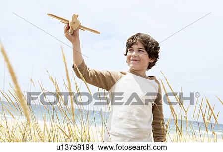 Picture - Smiling Pre-teen boy standing, arms raised on beach among plants, flying toy airplane. Fotosearch