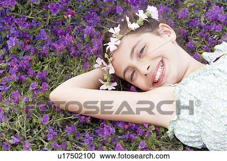 Stock Image - Smiling Pre-teen girl with flower headdress lying, arm behind head in field of flowers, elevated view. Fotosearch