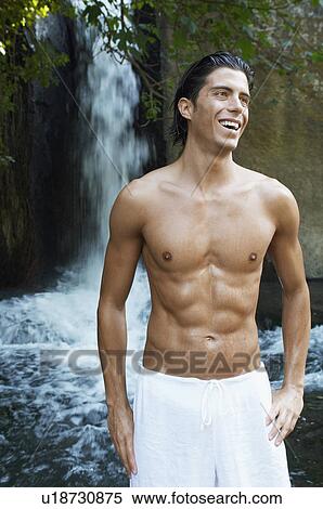 Stock Photography - Smiling young man standing by waterfall, front view. Fotosearch
