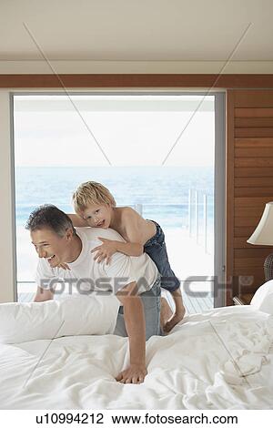 Stock Image - Son riding piggy back on father on bed. Fotosearch
