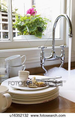 Stock Image - Stack of Dirty Dishes and silverware by Sink with running water below window with flower. Fotosearch