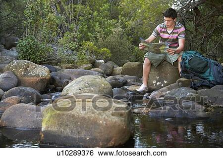 Stock Photograph - Teenage boy (16-17 years) sitting on stone by river reading map. Fotosearch