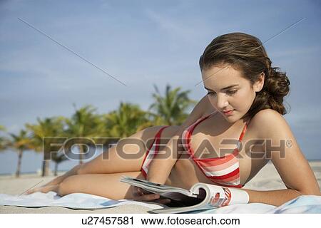 Stock Image - Teenage girl (16-17) reading magazine, lying on beach. Fotosearch