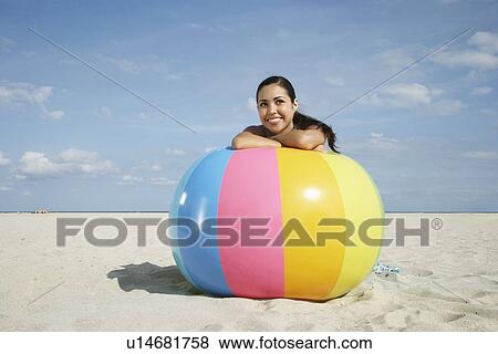 Stock Photo - Teenage girl (16-17) sitting behind beach ball on beach, portrait. Fotosearch