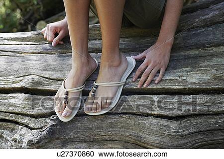 Stock Image - Teenage girl (16-17 years) wearing flip-flops sitting on tree trunk, low section. Fotosearch