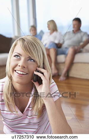 Stock Image - Teenage Girl sitting on floor, Talking on Cell Phone, family sitting in background. Fotosearch