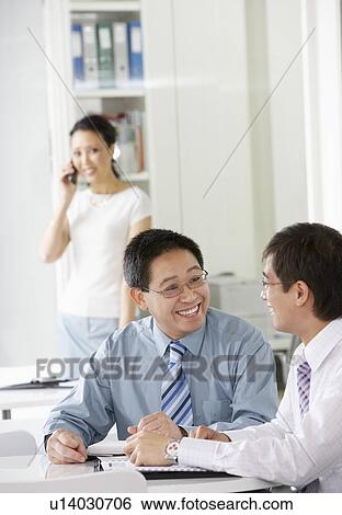 Stock Photograph - Two business man sitting at desk in office, woman on cell phone in background. Fotosearch