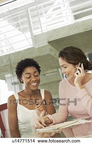 Picture - Two businesswomen in conference meeting, one using mobile phone. Fotosearch