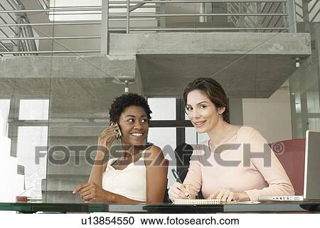 Stock Image - Two businesswomen in office, one using mobile phone, portrait. Fotosearch