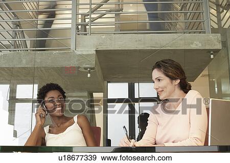 Stock Photo - Two businesswomen in office, one using mobile phone. Fotosearch