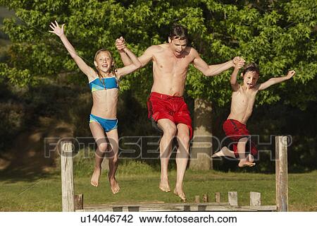 Stock Image - Two children (7-9) holding hands with father, jumping into lake.. Fotosearch