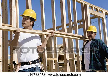 Stock Image - Two construction workers carrying lumber on construction site. Fotosearch