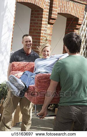 Stock Photo - Two men outside house carrying woman on sofa. Fotosearch