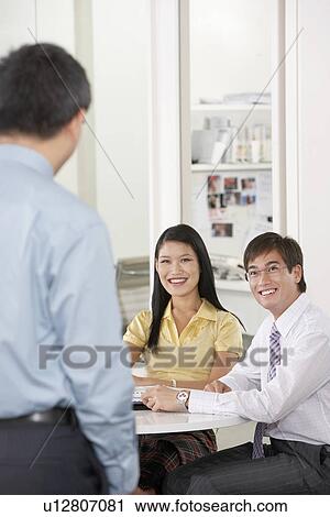 Two office workers sitting at table smiling at colleague View Large Photo Image Stock Image - Two office workers sitting at table smiling at colleague. Fotosearch
