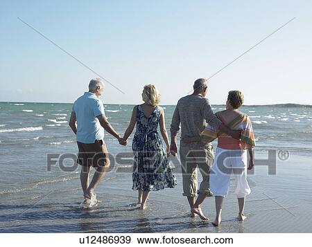 Stock Photo - Two senior couples walking on beach, back view. Fotosearch