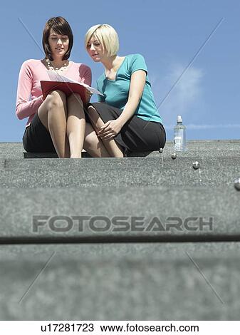 Stock Image - Two women sitting on stairs outdoors, reading papers, low angle view. Fotosearch