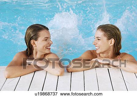Stock Photo - Two young women in pool at poolside splashing feet, front view. Fotosearch
