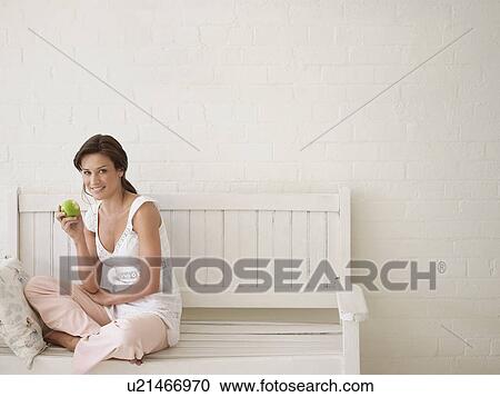 Stock Image - Woman Eating Apple, sitting, cross-legged on bench. Fotosearch
