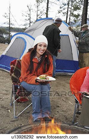 Stock Image - Woman eating sandwich by campfire, (portrait). Fotosearch