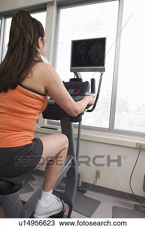 Stock Image - Woman Exercising on a Stationary Bike with a TV. Fotosearch