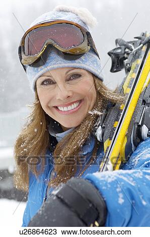 Stock Image - Woman holding skis in snow, head and shoulders, portrait.. Fotosearch