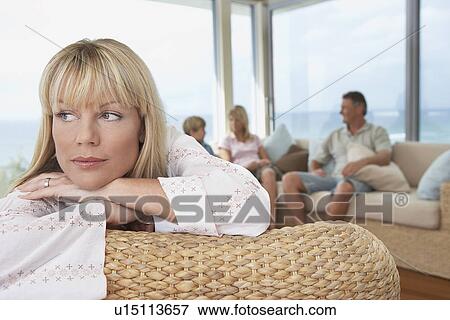 Stock Photo - Woman lying, arms crossed, over back of chair, family in background. Fotosearch