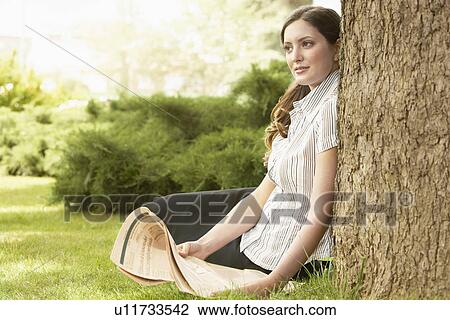 Woman sitting in grass, leaning on tree, holding newspaper. View Large Photo Image Stock Image - Woman sitting in grass, leaning on tree, holding newspaper.. Fotosearch