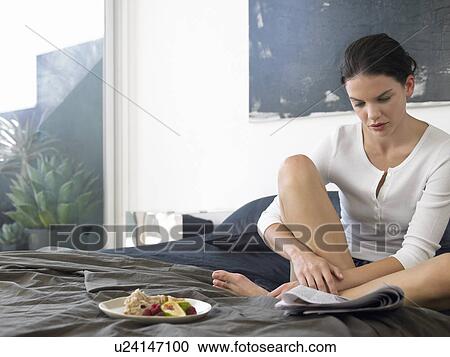 Stock Image - Woman sitting on bed with food, reading paper. Fotosearch