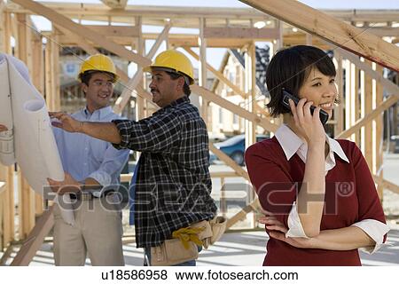 Stock Photo - Woman using mobile phone near workers on construction site. Fotosearch