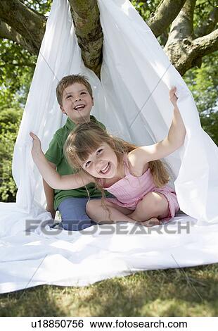 Stock Photograph - Young boy and girl in backyard sitting in tent made of bed sheet. Fotosearch