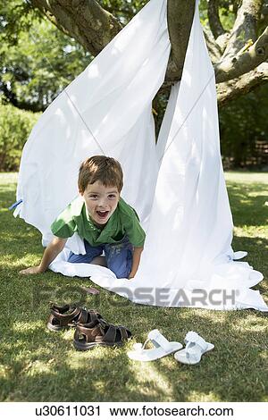 Stock Image - Young boy in back yard crawling out of tent made of bed sheet, portrait. Fotosearch