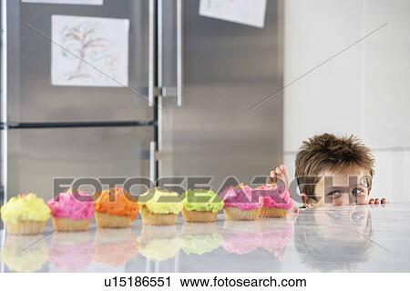 Stock Image - Young boy peaking over counter at row of cupcakes in kitchen. Fotosearch