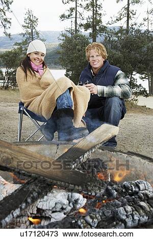 Stock Image - Young couple in warm clothing, sitting by campfire. Fotosearch