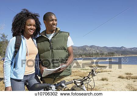 Stock Image - Young Couple on Mountain Bikes Using Map by lake. Fotosearch