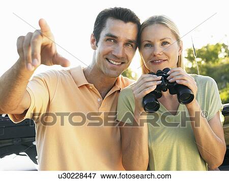 Young couple outdoors, man pointing, woman holding binoculars, smiling View Large Photo Image Stock Photo - Young couple outdoors, man pointing, woman holding binoculars, smiling. Fotosearch