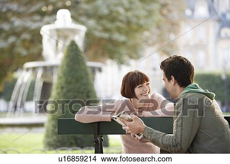 Picture - Young couple reading guide book, sitting on bench, in park. Fotosearch