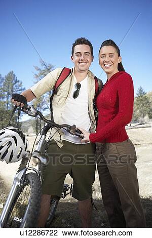 Stock Image - Young couple standing in field with mountain bike.. Fotosearch