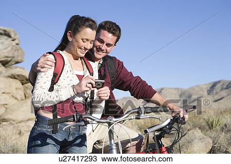 Stock Image - Young couple with mountain bikes stand looking at camera. Fotosearch