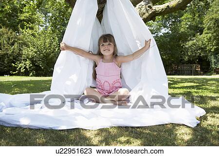 Stock Image - Young girl in backyard sitting in tent made of bed sheet. Fotosearch