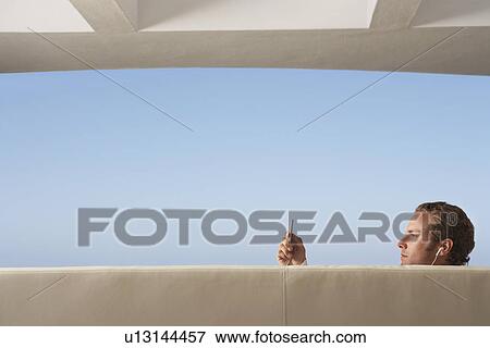 Stock Photo - Young man holding mp3 player listening to music through earphones, profile. Fotosearch
