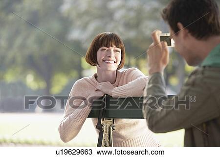Stock Image - Young man photographing young woman on bench in park. Fotosearch