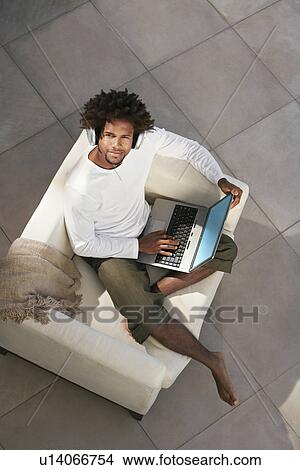 Picture - Young man sitting on sofa outside using laptop and listening to music through earphones, portrait, overhead view. Fotosearch