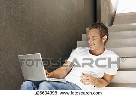 Stock Photo - Young man sitting on stairs using laptop, portrait. Fotosearch