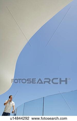 Stock Image - Young man using mobile phone on balcony, portrait, low angle view. Fotosearch